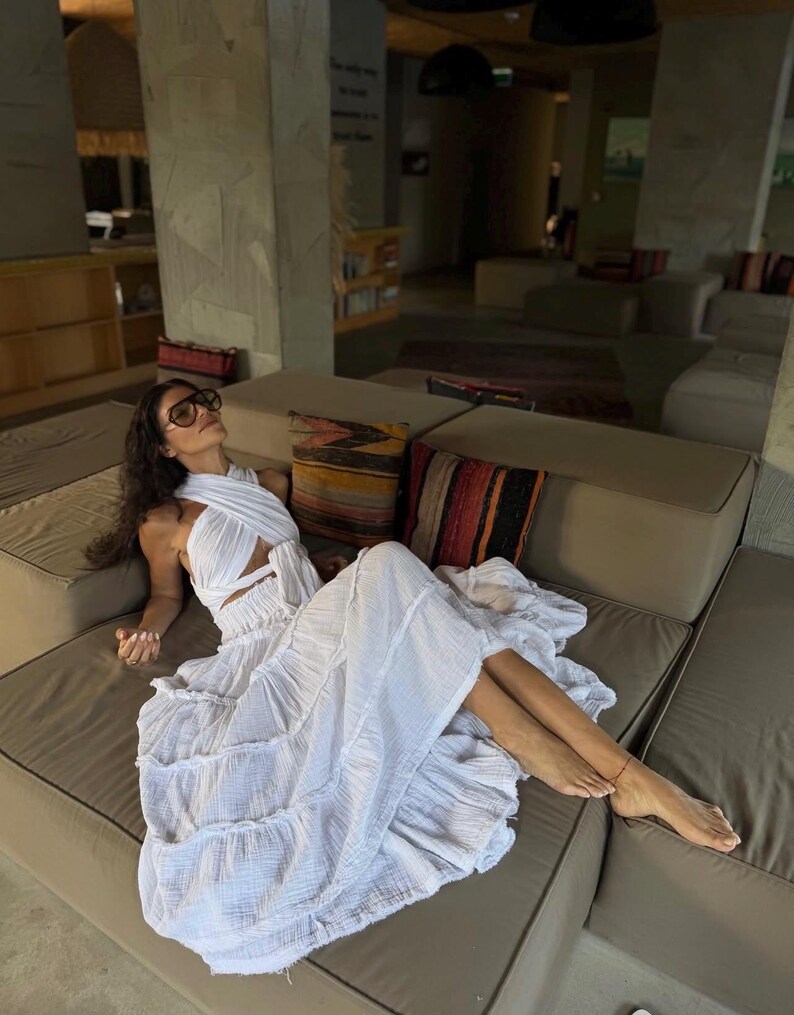 May include: A white, layered maxi dress with a halter neckline and cut-out detail. The dress features a tiered skirt and is worn with sunglasses. The person is lounging on a neutral-toned sofa with decorative pillows.Sacred Ceremony Wear.