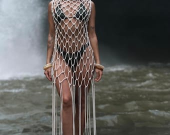 Könnte beinhalten: Eine Frau trägt ein weißes, gehäkeltes Maxikleid mit Fransen am Saum. Das Kleid ist netzartig und wird über einem schwarzen Bikini getragen. Sie steht auf Felsen in der Nähe eines Wasserfalls.