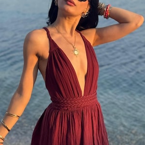May include: A deep burgundy maxi dress featuring a plunging V-neckline, thin braided shoulder straps, and a wide braided waistband. The garment is crafted from a textured, lightweight fabric with a gathered skirt that flows from the waist.