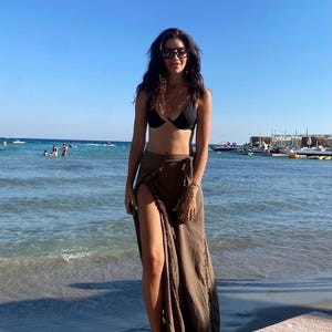 May include: A woman wearing a black bikini top and a brown wrap skirt stands on a sandy beach with blue water in the background.