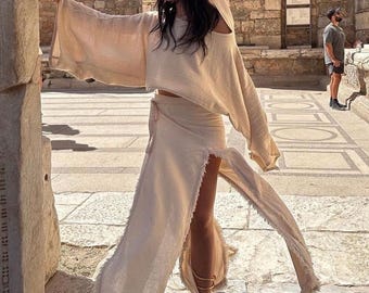 May include: A beige two-piece set consisting of a hooded long-sleeve crop top with wide sleeves and a matching wrap skirt featuring a high side slit and frayed raw edges along the hemline, crafted from a textured, lightweight fabric.