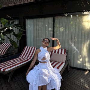 May include: A white two-piece set consisting of a halter-style crop top with a crisscross front design and a matching tiered midi skirt with a gathered waistband. The fabric has a textured appearance, and the outfit is paired with white sandals.