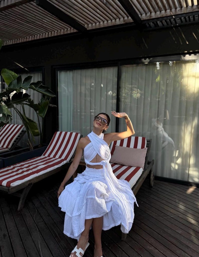 May include: A white two-piece set consisting of a halter-style crop top with a crisscross front design and a matching tiered midi skirt with a gathered waistband. The fabric has a textured appearance, and the outfit is paired with white sandals.