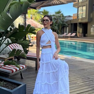 May include: A white two-piece set consisting of a sleeveless halter-style crop top with a crisscross neckline and a matching floor-length tiered maxi skirt with a gathered elastic waistband, featuring a textured fabric finish throughout the garment.
