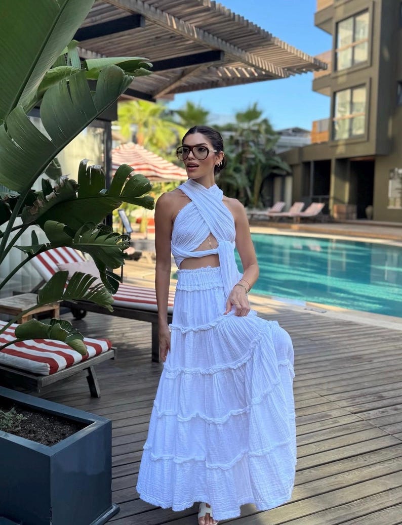 May include: A white two-piece set consisting of a sleeveless halter-style crop top with a crisscross neckline and a matching floor-length tiered maxi skirt with a gathered elastic waistband, featuring a textured fabric finish throughout the garment.