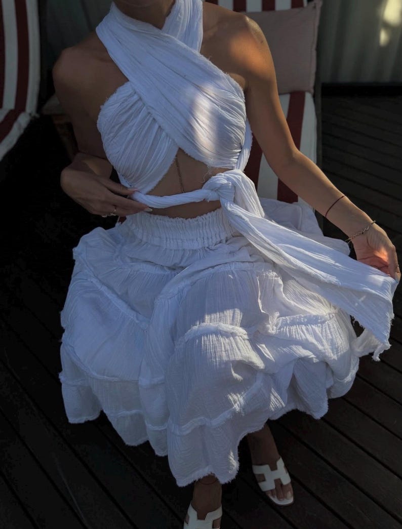 May include: A white two-piece set consisting of a halter-style crop top with a crisscross front design that ties at the waist and a matching tiered midi skirt with a gathered elastic waistband, both made from a textured, lightweight fabric.