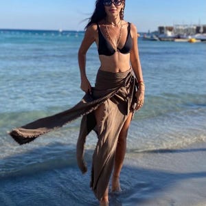 May include: A woman wearing a black bikini top and a brown wrap skirt stands on a sandy beach with the ocean in the background.