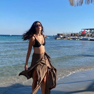 May include: A woman wearing a black bikini top and a brown wrap skirt with a high slit. She is standing on a sandy beach with the ocean in the background.