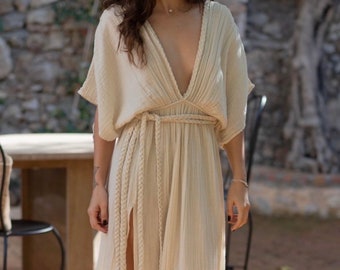 Puede incluir: Vestido largo color crema con escote en V profundo y mangas cortas. Cuenta con un cinturón trenzado y una abertura alta en el lado izquierdo. La tela tiene una apariencia texturizada. El vestido se usa al aire libre.