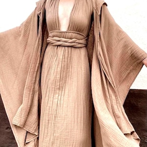 Puede incluir: Un vestido beige largo hasta el suelo con un escote en V profundo y una cintura trenzada, combinado con una capa a juego de manga larga con capucha. La prenda presenta un tejido texturizado y arrugado que crea una silueta drapeada.