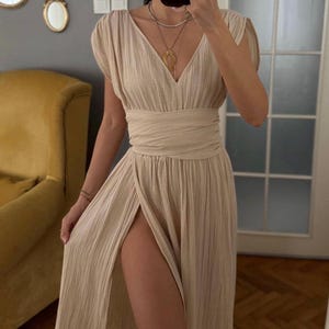 Puede incluir: Un vestido largo de color beige con escote en V, mangas cortas y una cintura ancha envuelta. La falda tiene una apariencia texturizada y plisada con una abertura frontal alta que se extiende desde la cintura hasta el dobladillo.