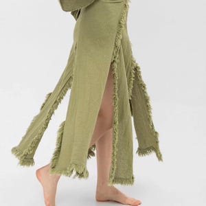 May include: A long, green skirt with a fringed hem and a wrap-style closure. The skirt has two slits on the sides, revealing the legs.