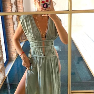 May include: A pale green maxi dress with a plunging neckline and a high split. The dress has a braided waist detail and a flowing, pleated design. The dress is sleeveless and appears to be made of a lightweight fabric.