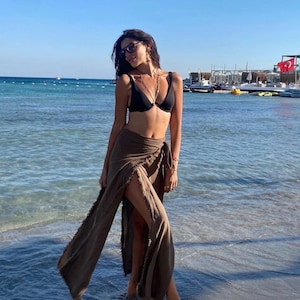 May include: A woman wearing a black bikini top and a brown wrap skirt with a slit up the side. She is standing on a sandy beach with the ocean in the background.