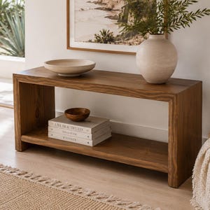 May include: A rectangular wooden console table with a lower shelf. The top surface holds a shallow beige bowl and a large, round, textured off-white vase containing green foliage. The lower shelf holds a small wooden bowl and three stacked books.