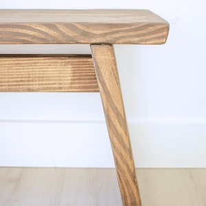 May include: A wooden bench featuring a flat rectangular seat and a single angled leg. The wood displays a visible grain pattern with a medium brown stain finish. A horizontal support beam connects the leg to the main structure of the bench.