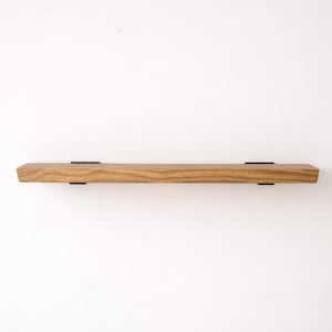 May include: A wooden floating shelf with a natural wood grain finish. The shelf is rectangular with a beveled edge and is supported by two black metal brackets. The shelf is designed for wall mounting and adds a rustic touch to any room.