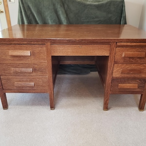 May include: A wooden desk with a warm, reddish-brown finish. The desk features three drawers on the left and right sides, with a central drawer above the knee space. The desk has a rectangular top and is supported by four legs.