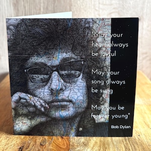 Può includere: Biglietto d'auguri con un ritratto di Bob Dylan con una sovrapposizione di mappa. Il biglietto include la citazione "May your heart always be joyful, May your song always be sung, May you be forever young" di Bob Dylan.