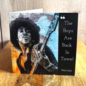 May include: Greeting card featuring a portrait of a musician playing a bass guitar, set against a map background. The card has the text "The Boys Are Back In Town!" and the band name "Thin Lizzy" on a black background.