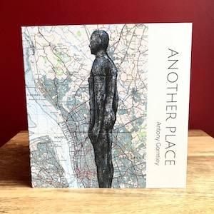 Puede incluir: Una imagen en blanco y negro de una escultura de pie sobre un mapa de Liverpool, Inglaterra. La escultura es una figura humana, y el mapa muestra las calles y vías fluviales de la ciudad. El texto "Another Place" y "Antony Gormley" está impreso en el lado derecho de la imagen.