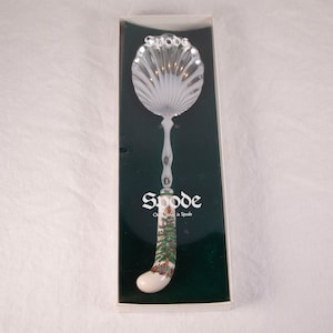 May include: A silver spoon with a white ceramic handle featuring a Christmas tree design. The handle is decorated with a green and red Christmas tree and the text "Spode Christmas in Spode".