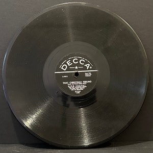 Op de afbeelding: Zwarte vinylplaat met een wit label. Het label heeft de tekst "Decca" in een witte cirkel met een ster in het midden. De tekst "That Christmas Feeling" staat onder het Decca-logo, gevolgd door "Bing Crosby" en "with the John Scott Trotter Orchestra and His Singers". Het platennummer "27229" staat onderaan het label.