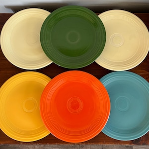 May include: Six colorful ceramic plates in shades of yellow, green, orange, and blue. Each plate has a concentric ring design and a central circular indentation. The plates are arranged on a dark wooden surface.