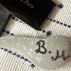 Puede incluir: Un cepillo para el cabello gris con un diseño de diamantes de imitación negros que deletrea "3.1..."