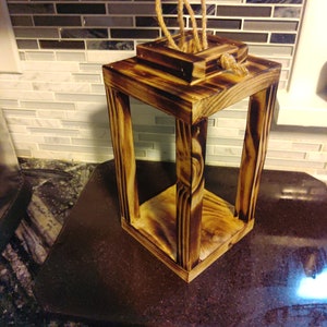 May include: A rustic wooden lantern with a square shape and a rope handle. The lantern has a dark brown wood finish with a burnt wood effect.