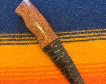 May include: A handcrafted knife with a dark, textured blade and a patterned wooden handle. The handle is a light brown color, and the blade is a dark, almost black color. The knife is set against a backdrop of blue, orange, and yellow striped fabric.