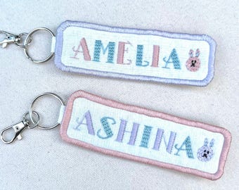 May include: Two embroidered keychains with names. One keychain has the name "AMELIA" in pink and blue letters, and the other has the name "ASHINA" in blue and purple letters. Each keychain has a small embroidered bunny and a silver clasp.