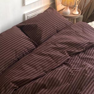 Puede incluir: Una cama con una funda nórdica a rayas burdeos y fundas de almohada a juego. La ropa de cama está cuidadosamente colocada sobre la cama, con un aspecto suave y acogedor. La habitación tiene una paleta de colores neutros, con una pared blanca y una mesita auxiliar con una lámpara.