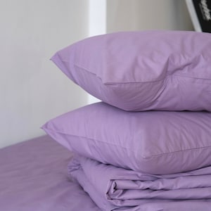 May include: A stack of three lavender-colored pillowcases and a folded lavender-colored duvet cover. The duvet cover is folded in thirds and the pillowcases are stacked on top of it. The bedding is on a lavender-colored bed with the text "bavéna HOME" printed on the bottom right corner.