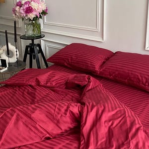 Op de afbeelding: Een bed met een rood gestreept dekbedovertrek en bijpassende kussenslopen. Het beddengoed is netjes opgemaakt, met het dekbed gedeeltelijk teruggeslagen. Een boeket roze en witte bloemen in een glazen vaas staat op een kleine zwarte tafel.