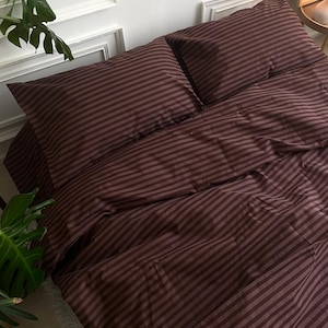 May include: A burgundy striped bedding set, including two pillowcases and a duvet cover, is neatly arranged on a bed. The fabric has a soft sheen, and the stripes run horizontally across the surface. The overall aesthetic is simple and elegant.