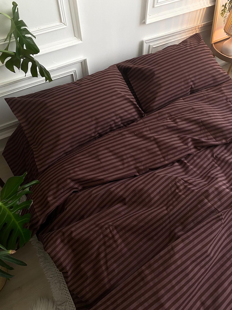 May include: A burgundy striped bedding set, including two pillowcases and a duvet cover, is neatly arranged on a bed. The fabric has a soft sheen, and the stripes run horizontally across the surface. The overall aesthetic is simple and elegant.