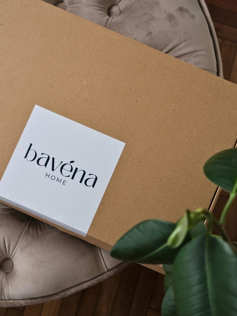 May include: A large, unopened cardboard box with a white label that reads "bav&eacute;na HOME" in black text. The box is resting on a tufted, beige upholstered surface. Green plant leaves are visible in the lower right corner.