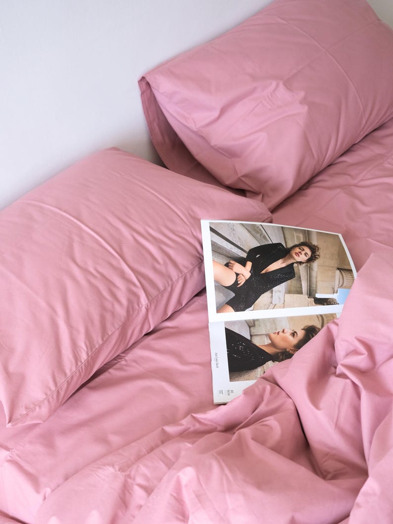 May include: A pink bed with two pillows and a magazine on top of the bedding. The magazine features a woman wearing a black outfit.