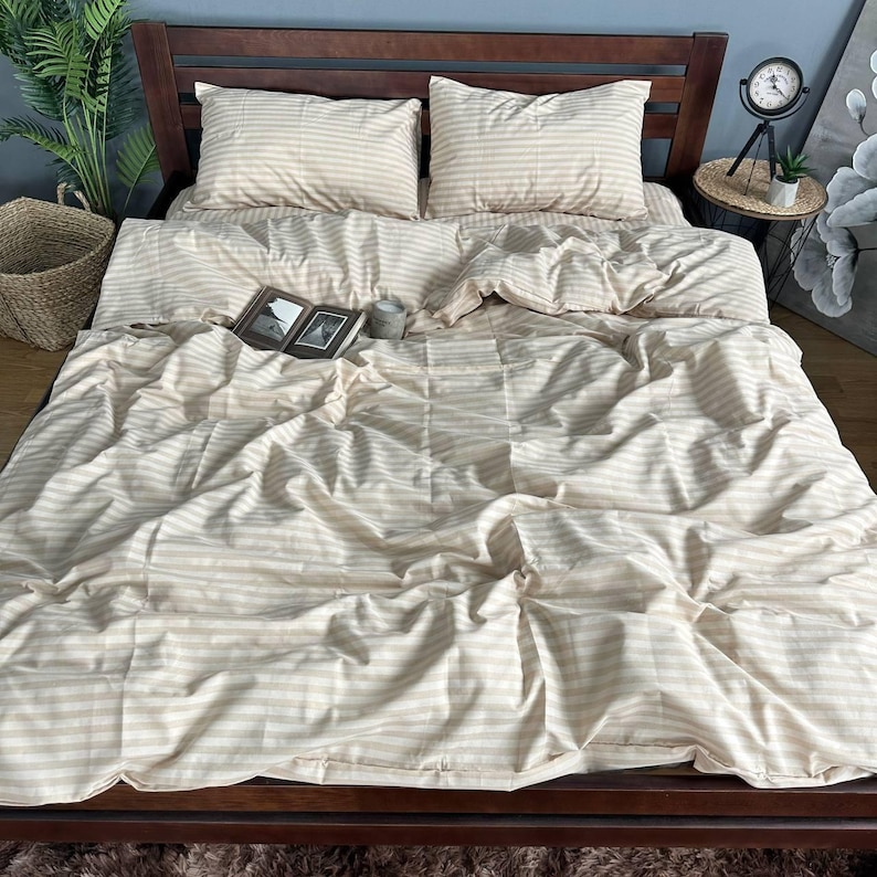 May include: A bed with a striped beige and white comforter set. Two matching pillowcases are visible, along with a book and a small cup. The bed frame is dark wood.