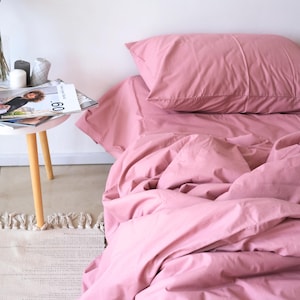 May include: A dusty rose colored duvet cover and two matching pillowcases are on a bed. The duvet cover is slightly rumpled and the pillowcases are smooth. The bed is made and the bedding is neatly arranged.