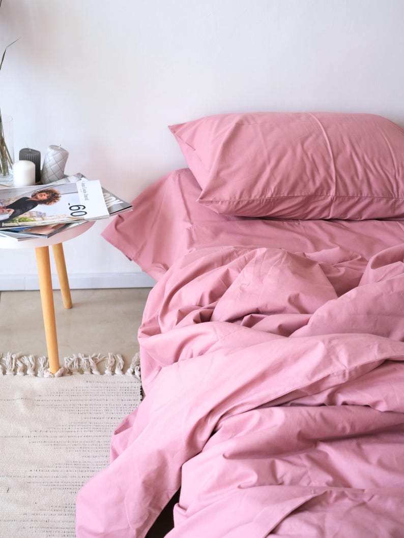 May include: A dusty rose colored duvet cover and two matching pillowcases are on a bed. The duvet cover is slightly rumpled and the pillowcases are smooth. The bed is made and the bedding is neatly arranged.