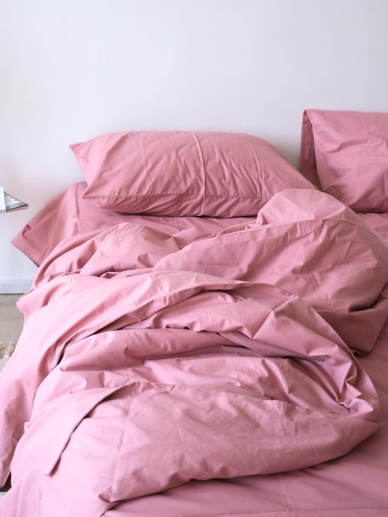 May include: A pink bed with a white headboard and two pink pillows. The bed is made with a pink duvet cover and the duvet is slightly rumpled.