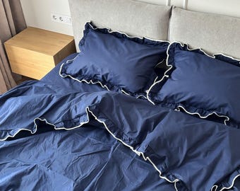 Può includere: Un set di biancheria da letto blu navy profondo composto da un copripiumino e due federe abbinate. Ogni pezzo è rifinito con un delicato bordo a volant bianco, che crea un netto contrasto con il tessuto blu scuro a tinta unita.
