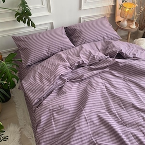 May include: A lavender and white striped bed set, including two pillowcases and a fitted sheet, is neatly arranged on a bed. The bedding is made of a soft, possibly cotton, material. The room has white walls and a decorative lamp.