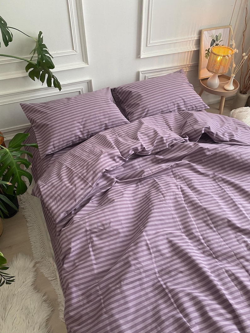 May include: A lavender and white striped bed set, including two pillowcases and a fitted sheet, is neatly arranged on a bed. The bedding is made of a soft, possibly cotton, material. The room has white walls and a decorative lamp.