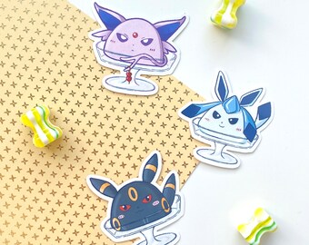 May include: Three Pokemon character stickers on a gold patterned background. The stickers feature the characters Gardevoir, Glaceon, and Umbreon, each in a dessert-like setting.