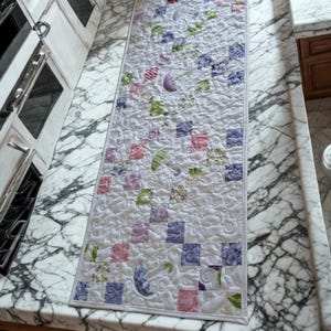 May include: A rectangular quilted table runner with a white background. The runner features a patchwork design with squares of pink, purple, and blue, along with floral and crescent moon motifs. The runner is approximately 60 inches long.