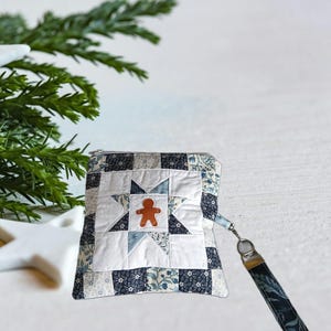 Puede incluir: Un estuche acolchado con un diseño de estrella y una aplicación de hombre de jengibre. El estuche tiene un cierre de cremallera y una correa de muñeca con un estampado floral. La combinación de colores incluye blanco, azul y marino.