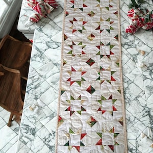May include: A rectangular quilted table runner featuring a repeating pattern of white, red, and green star blocks. The fabric is stitched with swirling patterns and finished with a light tan border. The design consists of alternating star motifs.